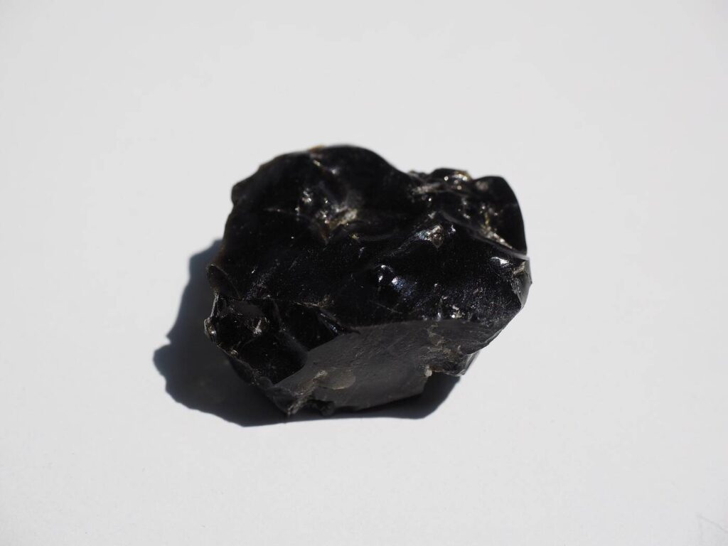 Schwarzer Obsidian Rohstein mit glasigem Glanz und scharfen Bruchkanten auf hellem Hintergrund